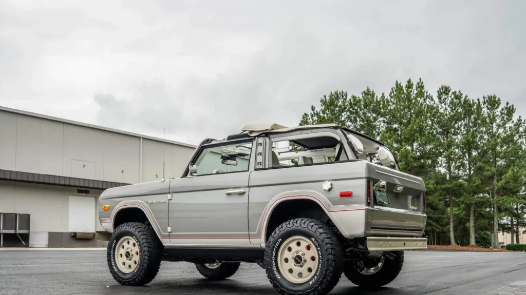 A vintage silver off-road vehicle with a weathered body, rugged tires, and an open roof parked on an asphalt lot near a warehouse and trees.