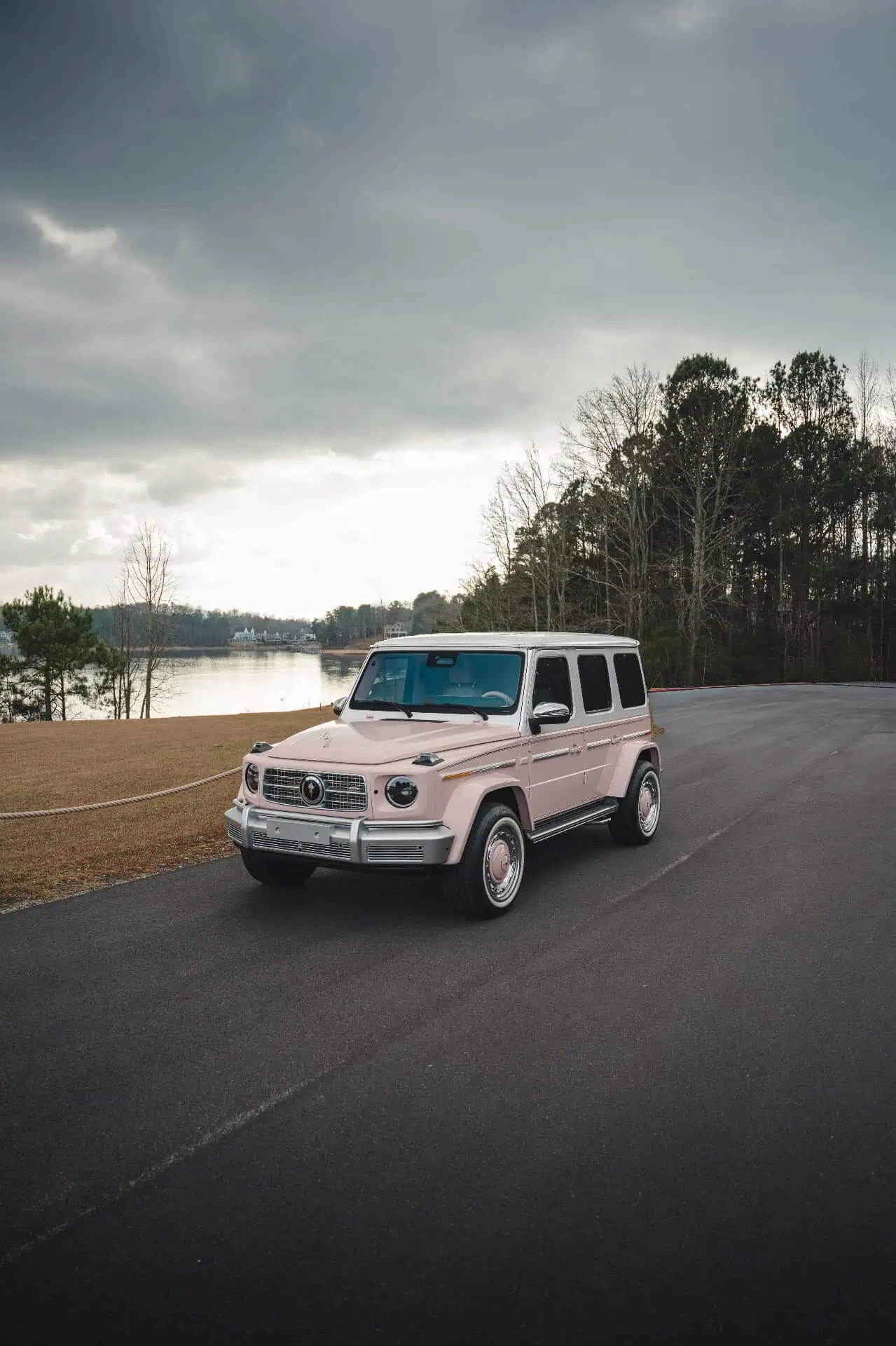 A pink vintage Mercedes-Benz G-Class SUV parked on a scenic road near a lake, with a dramatic cloudy sky and trees in the background, showcasing classic car style and outdoor elegance.